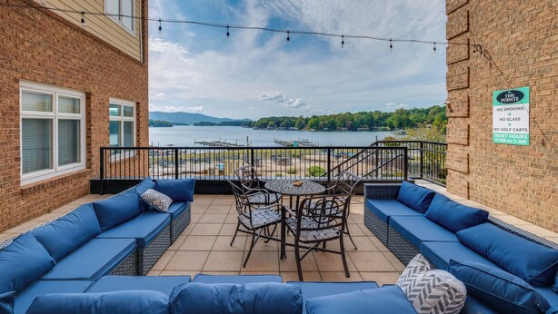 Un balcon avec des sièges bleus, une table et des chaises, donnant sur un lac où l’on peut voir des bateaux et un quai.