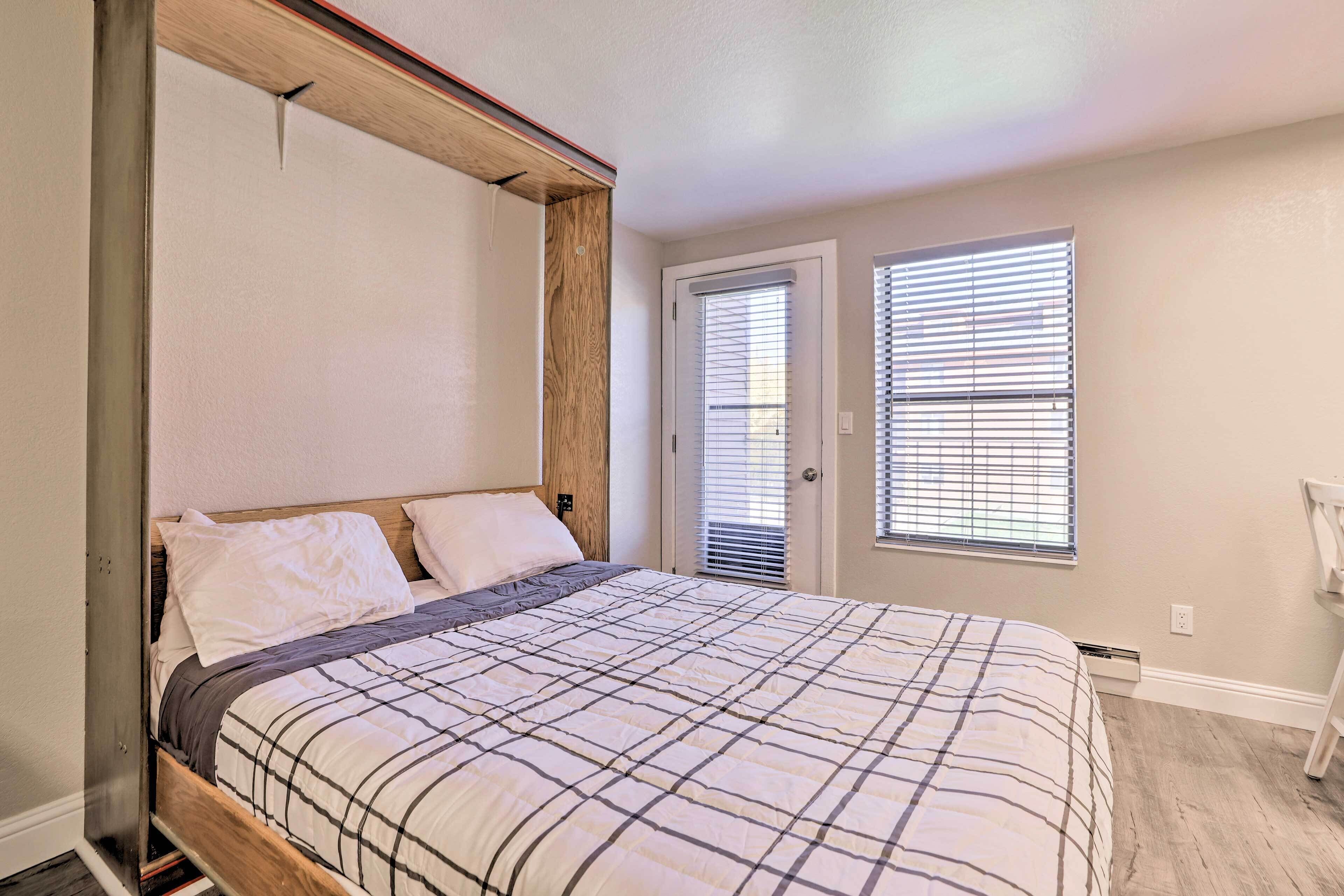 Un dormitorio con cabecera de madera, cama con colcha a cuadros, dos ventanas con persianas y una puerta.