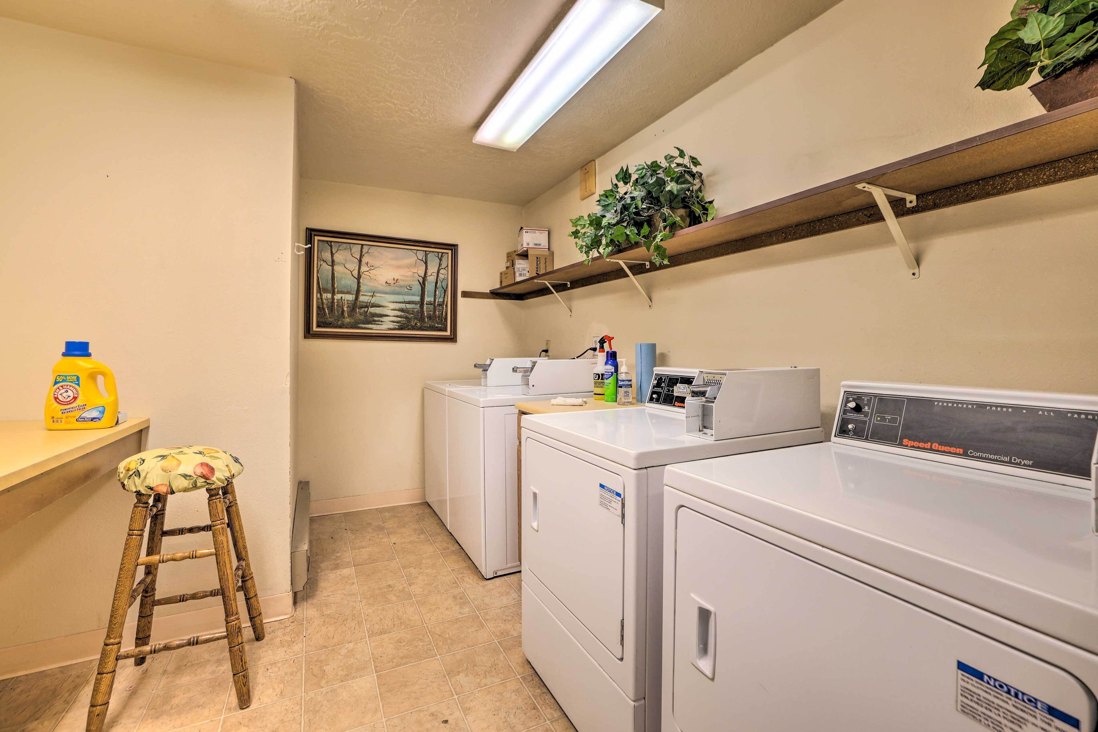 A laundry room with two washing machines, a dryer, a stool, a shelf with plants, and a painting on the wall.