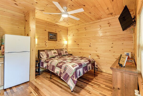Una habitación tipo cabaña acogedora con una cama, un refrigerador blanco, un ventilador de techo y un televisor montado.