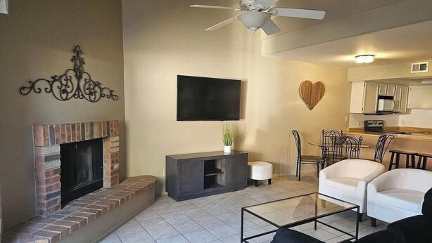 A living room with a fireplace, a flat-screen TV, a ceiling fan, and a kitchen area with white cabinets and a heart-shaped decoration. - NEW LOW RATE FOR THE SUMMER!                3-bed 2-bath townhome in Tempe (Tempe)