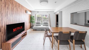 A modern living room with a flat-screen TV displaying 'NETFLIX', a wooden dining table with chairs, a sofa, and a large window with a view of buildings. - OTARU SAMPO 2 (Otaru)