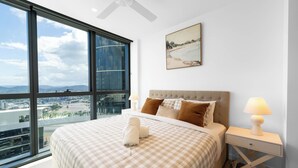 A bedroom with a large window, a bed with checkered bedding, a headboard, two bedside lamps, and a painting on the wall.