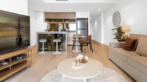 A modern living room with a flat-screen TV, a round coffee table with decorative items, a sofa, and a dining area with a kitchen in the background.