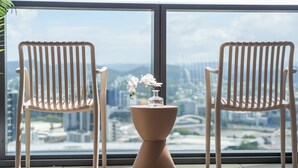 Two white chairs with a vase of white flowers on a pedestal table, positioned in front of a large window with a cityscape view.