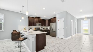 A modern kitchen with dark wooden cabinets, a white island with bar stools, stainless steel appliances, and a large window providing natural light. - Super Mario & Frozen Castle Ultimate Stay with Free Access to Resort by Disney (Kissimmee)