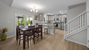 A modern kitchen with a central island, stainless steel appliances, and a dining area with wooden chairs. - Modern 3BR Home w/ Backyard, Firepit & Near DT (New Bern)