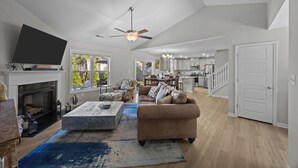 A spacious living room with a fireplace, a large TV, and a ceiling fan. - Modern 3BR Home w/ Backyard, Firepit & Near DT (New Bern)
