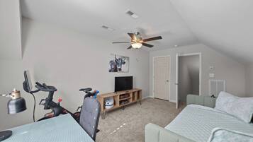 Habitación compacta y bien iluminada, con ventilador de techo, televisión, bicicleta y un pequeño rincón de descanso.