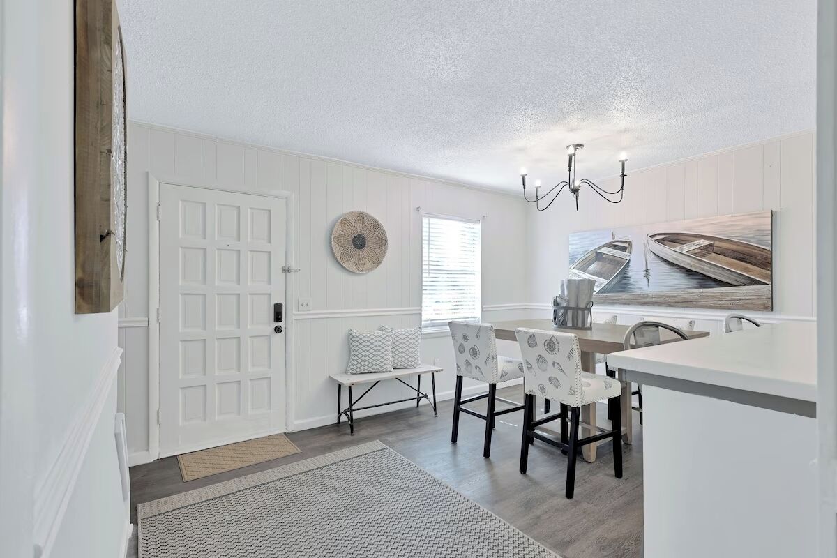 A dining area with a white table and chairs, a bench, a chandelier, and a wall-mounted artwork of a boat.