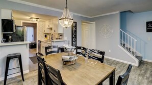 A modern kitchen with a chandelier, a dining area with a wooden table and chairs, and a staircase with a blue wall. - Hilton Head 2BR Escape w/ Pool & Sofa Bed (Hilton Head Island)