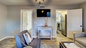 A living room with a grey sofa, a coffee table, and a TV mounted on the wall. There is a door leading to the kitchen in the background. - Hilton Head 2BR Escape w/ Pool & Sofa Bed (Hilton Head Island)