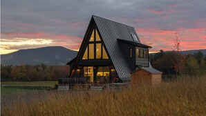 A modern house with a steeply pitched roof and large windows, surrounded by a field and mountains in the background. - Stowe A-Frame, Sauna, Hot Tub, Theater, Game Room, Arcade, Gym (Morristown)