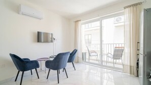 Ένα μοντέρνο σαλόνι με τηλεόραση επίπεδης οθόνης, δύο μπλε πολυθρόνες, ένα μικρό τραπέζι και μια συρόμενη γυάλινη πόρτα που οδηγεί σε μια εξωτερική βεράντα με καθίσματα.