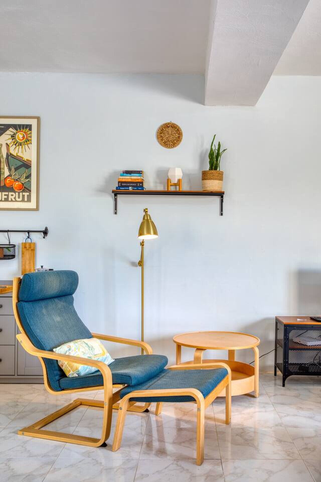 Ein Wohnzimmer mit blauem Sessel, einem kleinen Holztisch, einer Stehlampe und einem Bücherregal mit Büchern und einer Pflanze.