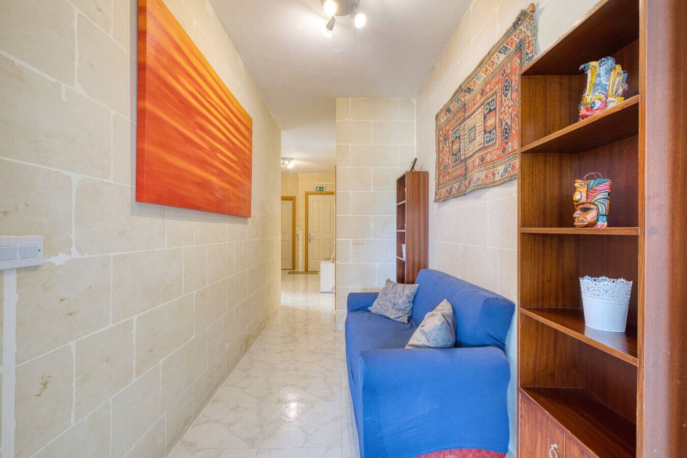 Un couloir avec un canapé bleu, une étagère en bois ornée d’objets décoratifs et une tapisserie colorée accrochée au mur.