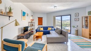 Ein geräumiges Wohnzimmer mit einem blauen Sofa, einem Holztisch und einem blauen Sessel. Ein Bücherregal mit Dekorationselementen und ein großes Fenster mit Blick ins Freie.