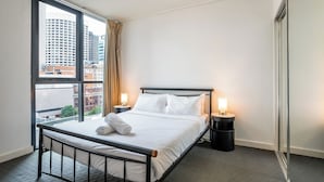 Une chambre d’hôtel avec une grande fenêtre, un lit, deux tables de chevet avec des lampes et une vue sur les immeubles.