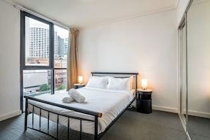 Habitación de hotel con un ventanal grande, una cama, dos mesitas de noche con lámparas y vistas a edificios.