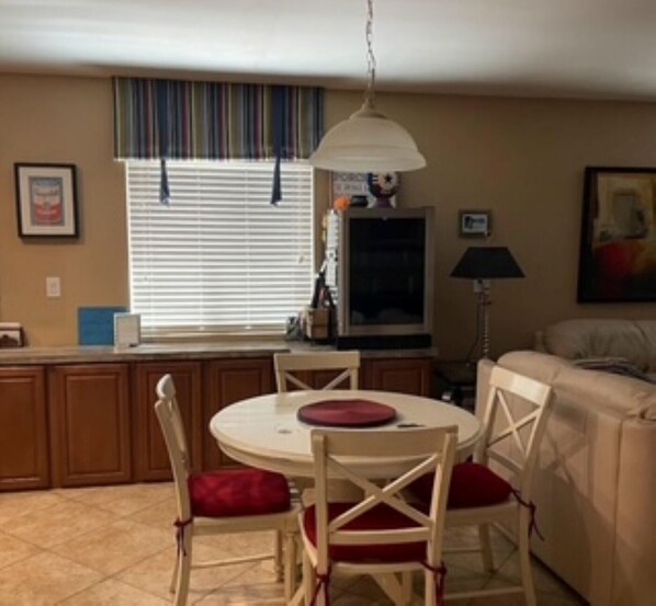 A dining area with a round table, four chairs, a hanging light fixture, a window with blinds, and a television on a stand.