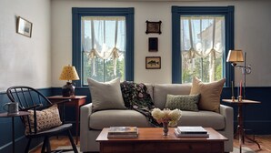 Una sala de estar acogedora con un sofá, una mesa de centro y una silla. Hay dos ventanas con detalles azules, una lámpara y un florero con flores sobre la mesa de centro.