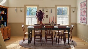 Comedor con mesa de madera, seis sillas y un jarrón de flores. Hay tres ventanas con cortinas de encaje y un aparador de madera con vajilla a la izquierda.
