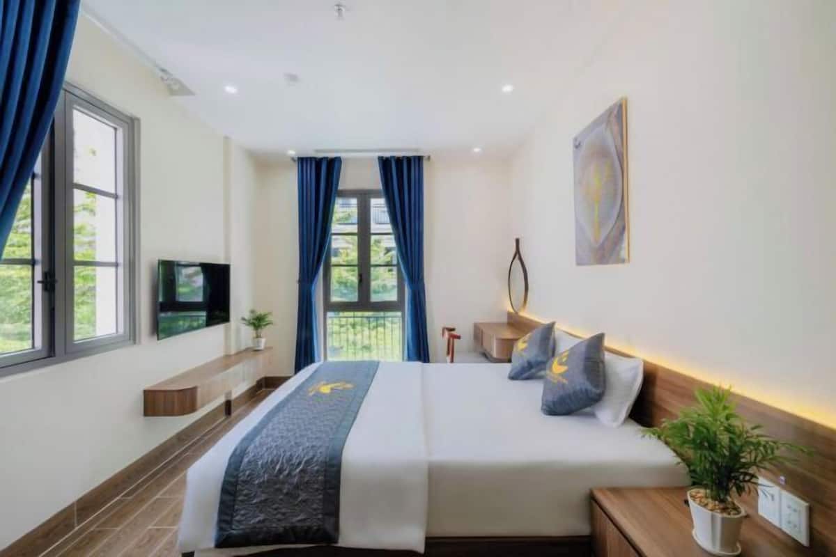 A modern hotel room with a large bed, a TV, a bedside table with a lamp, and a window with curtains.