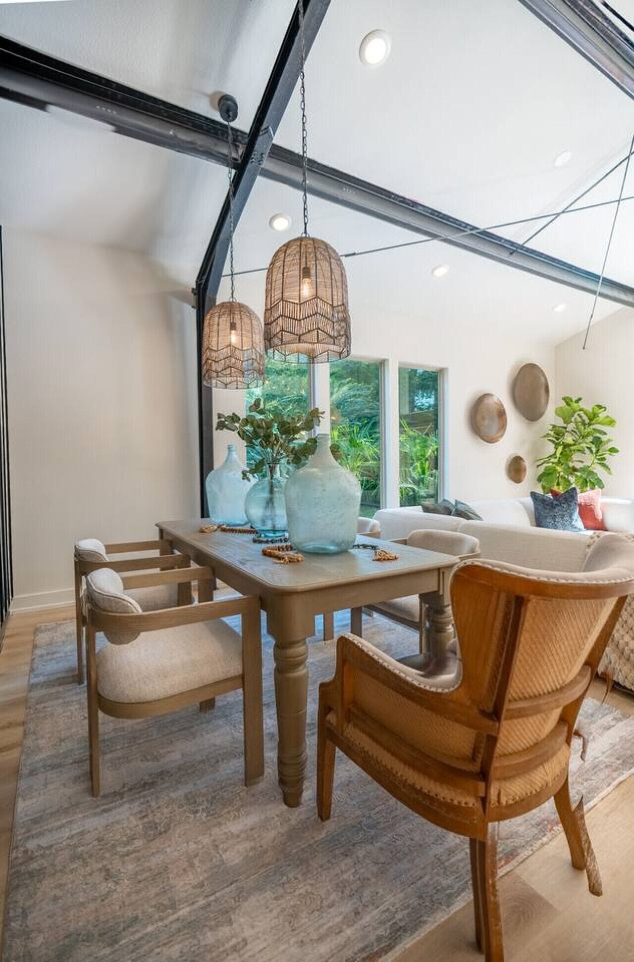 Un comedor con mesa de madera, sillas, lámparas colgantes y ventanas amplias que dan a vistas de vegetación exterior.
