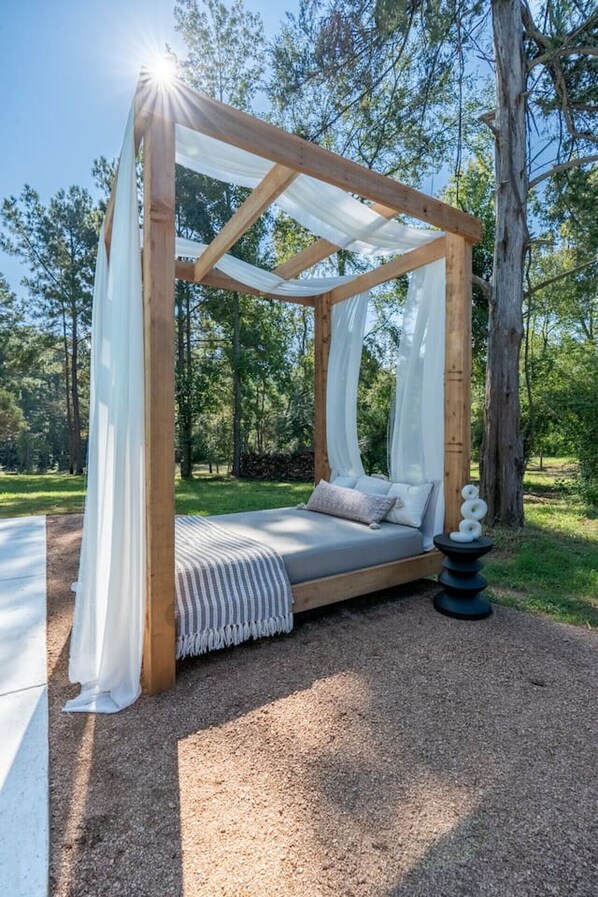 A wooden canopy bed with white drapes, set on a gravel path in a wooded area.