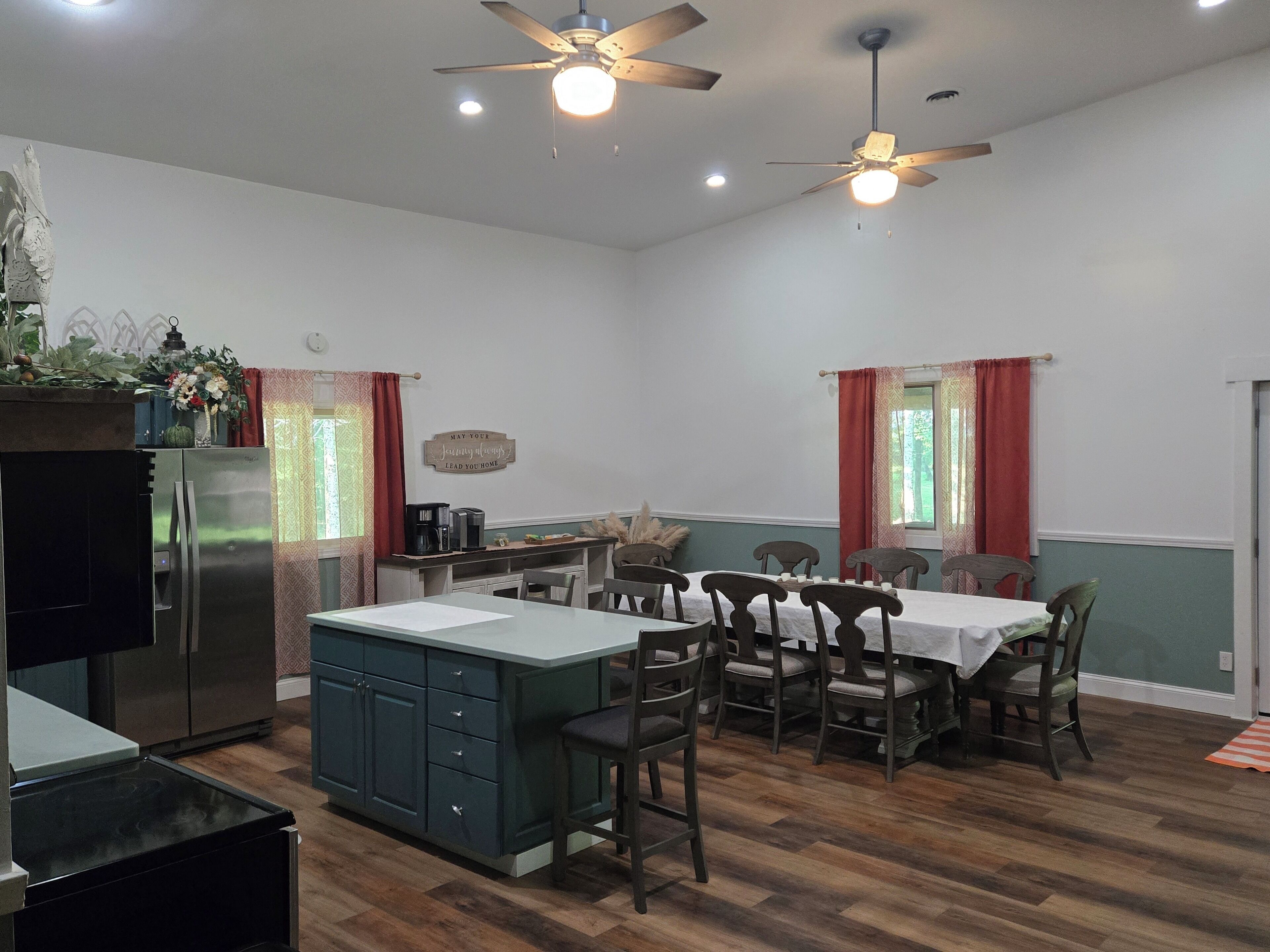 A spacious kitchen with a central island, wooden dining table, and chairs. There are ceiling fans and recessed lighting.