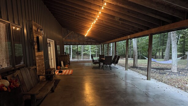 A covered porch with a concrete floor, wooden benches, a table, and chairs. There are string lights hanging from the ceiling.
