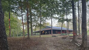 A rustic cabin with a covered porch and a fire pit area, surrounded by a forested area with fallen leaves on the ground.