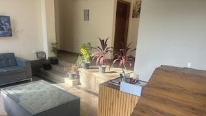A modern living room with a grey sofa, a glass-topped ottoman, and a wooden reception desk with a whiteboard. There are potted plants on the floor and a TV mounted on the wall. - Rc Stays (Noida)