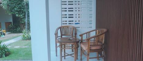 A balcony with two wooden chairs and a small table, a white partition, and a view of a garden.