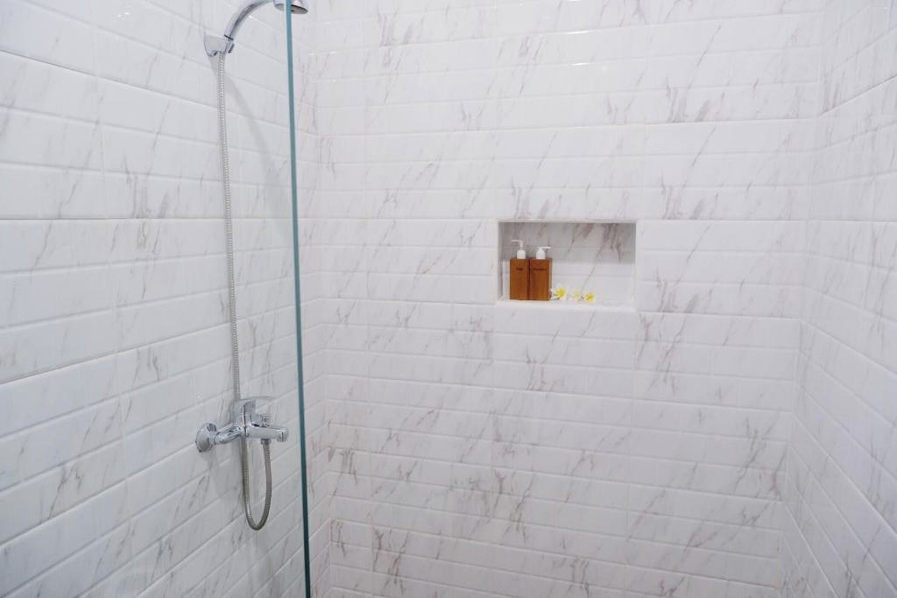 Douchegedeelte met witte marmeren tegels, een glazen deur en een klein plankje met toiletartikelen.
