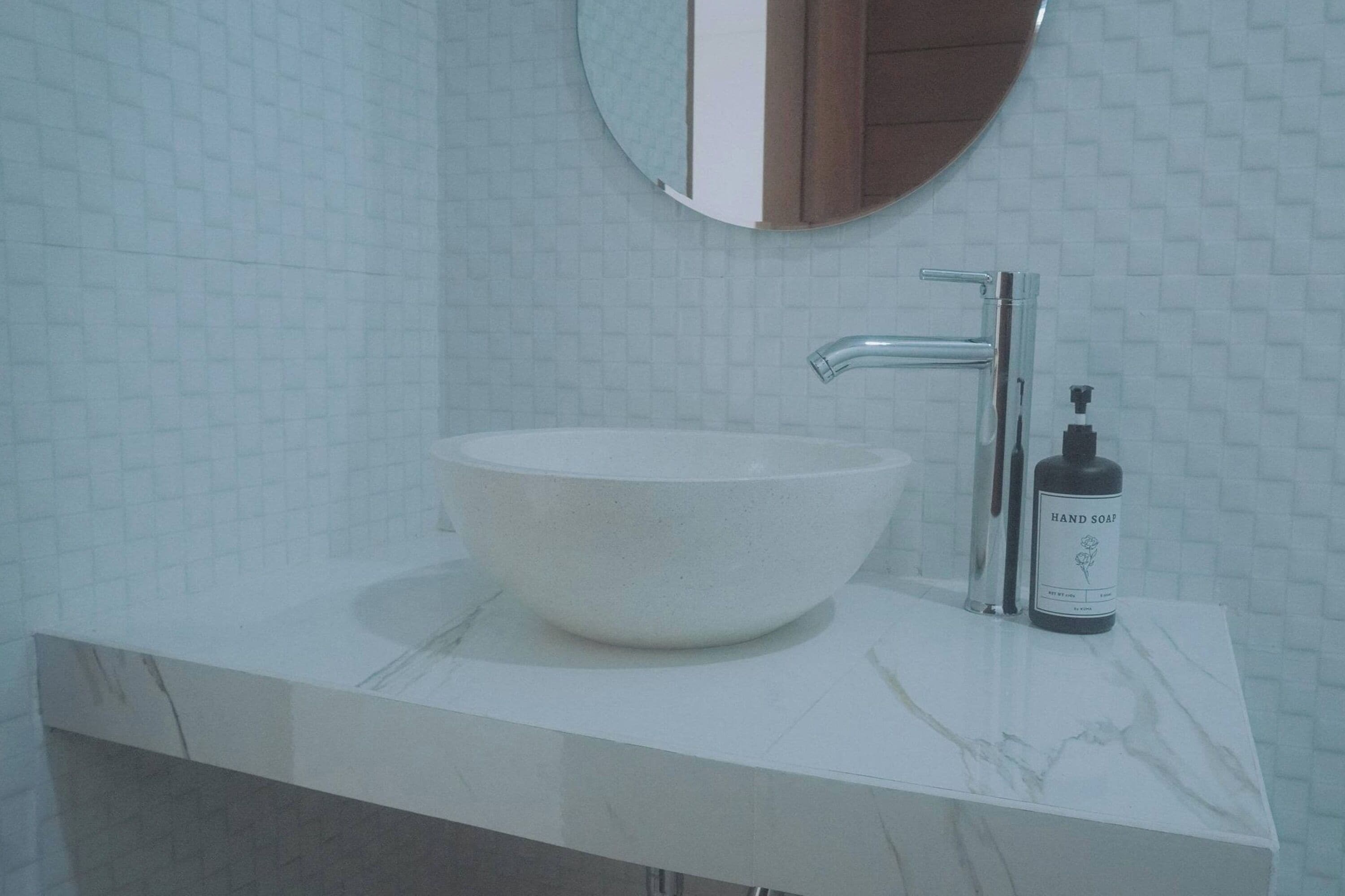 Un baño con un lavamanos de mármol blanco, un espejo redondo y una botella de jabón para manos.