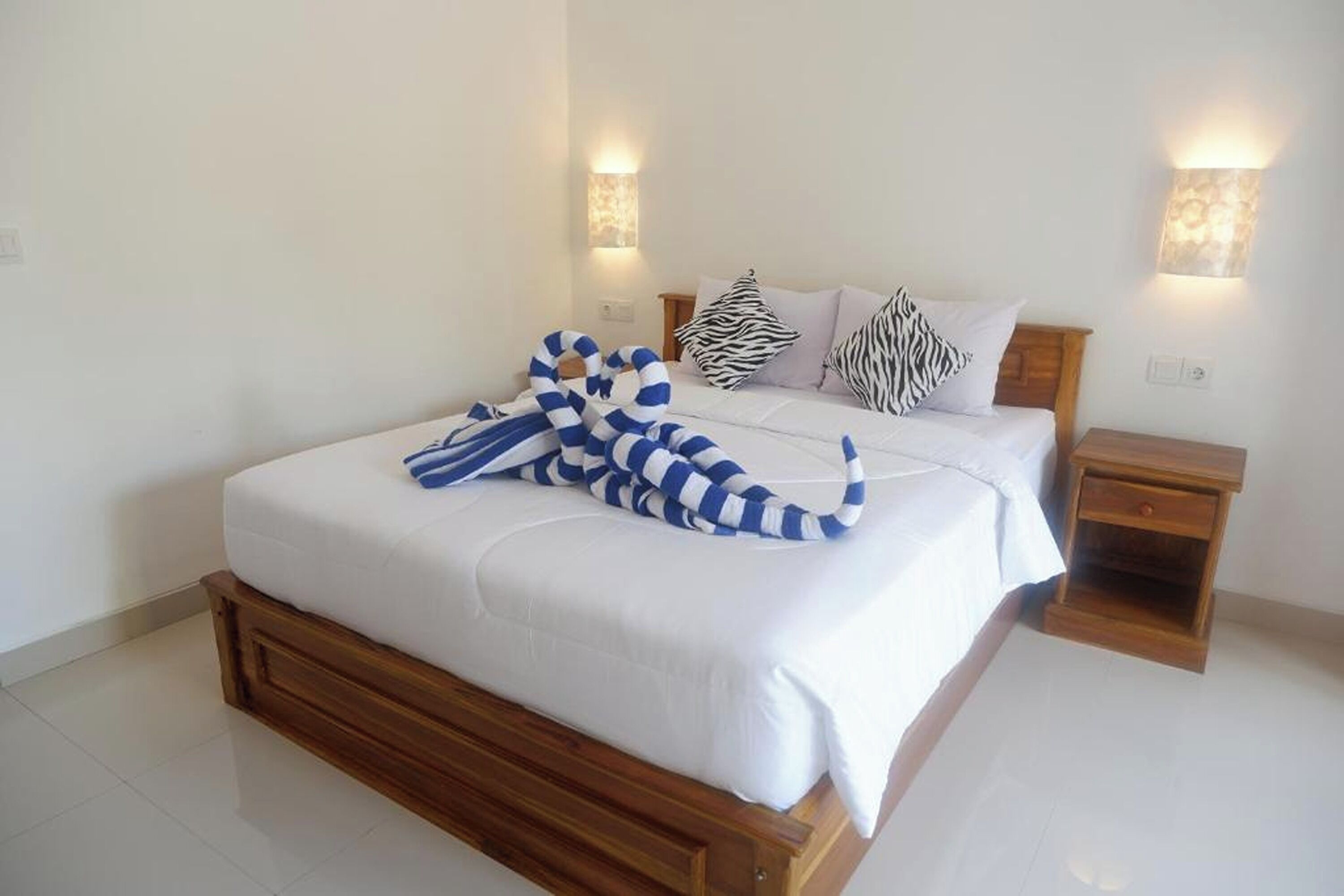 A neatly made bed with white bedding and striped pillows, a wooden nightstand, and wall-mounted sconces.