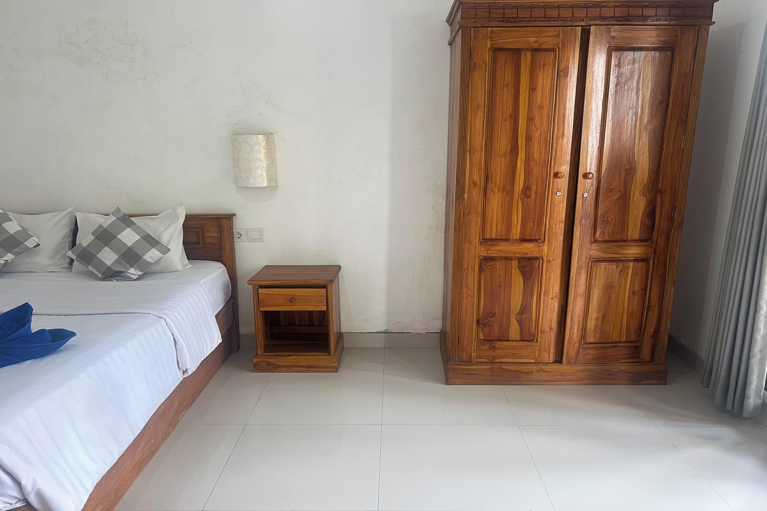 A bedroom with a wooden wardrobe, a bed with white bedding, a small wooden nightstand, and a wall-mounted light fixture.