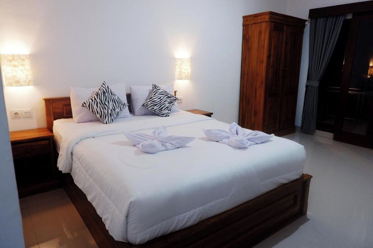 A neatly made bed with white linens and zebra-patterned pillows, a wooden wardrobe, and two wall-mounted lamps.