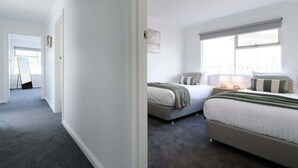 A bedroom with two beds, a window with blinds, a bedside table with a lamp, and a door leading to another room. - Modern Ease in East Devonport (City of Devonport)