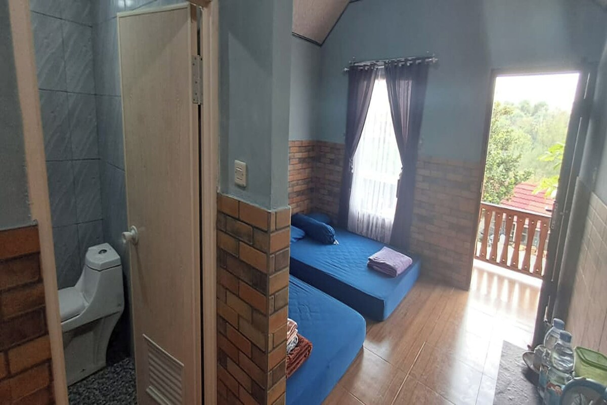 Ein Zimmer mit Toilette, einem Bett mit blauer Matratze und einem Fenster mit Vorhängen.