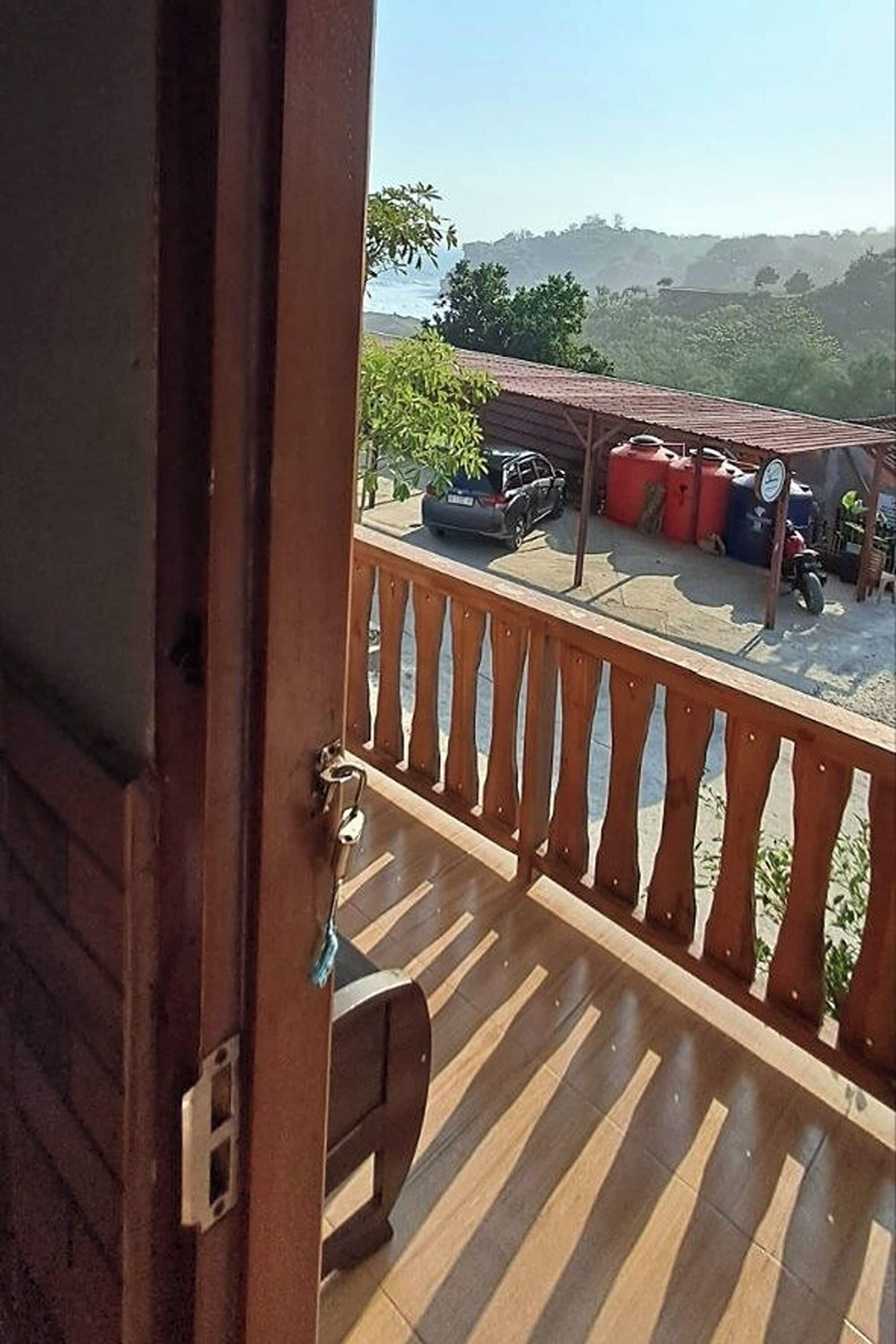Ein hölzerner Balkon mit Blick auf ein parkendes Auto und ein Motorrad, einen roten Container und einen wolkenlosen Himmel.