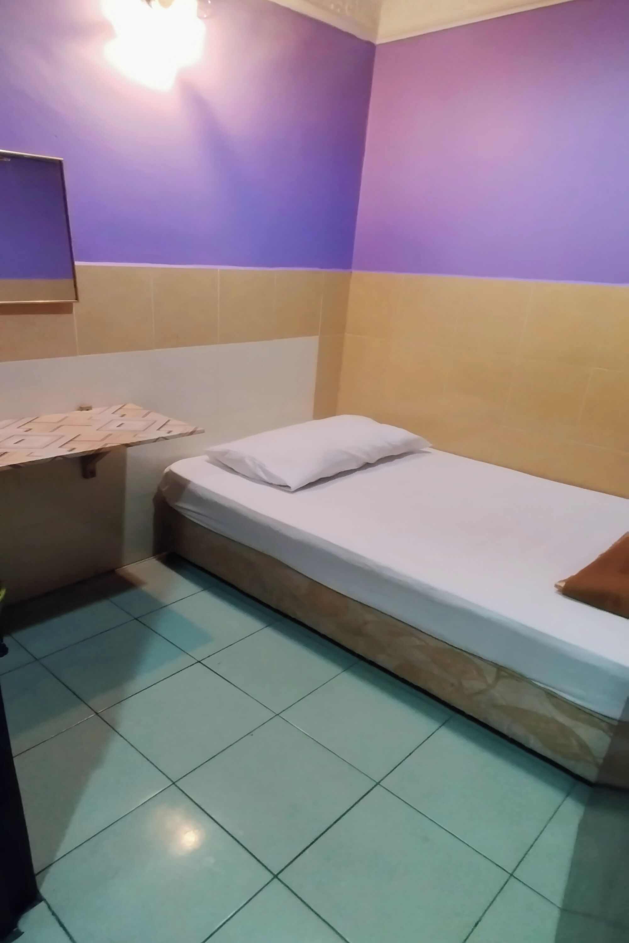 A small, single-bed room with a white mattress and pillow, a wooden headboard, and a tiled floor.