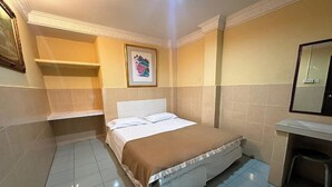 Een hotelkamer met een bed, een klein bureau, een stoel, een ingelijst beeld aan de muur en een plank boven het bed.