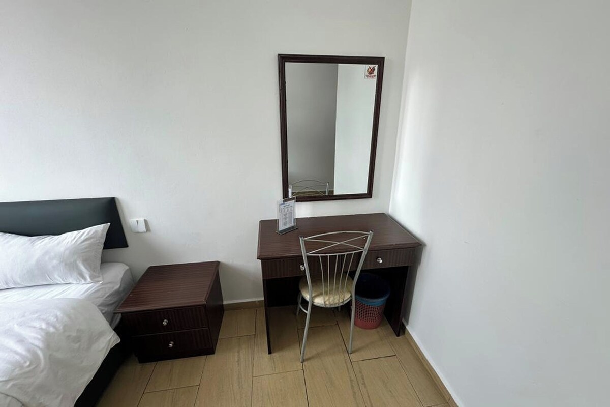 A hotel room with a bed, a desk, a chair, a mirror, and a trash bin.