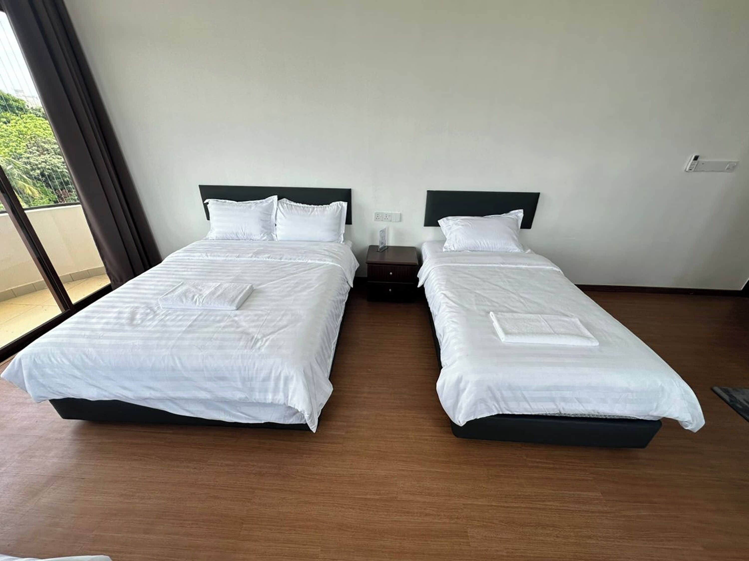 A hotel room with two single beds, a nightstand, and a window with a view of greenery.