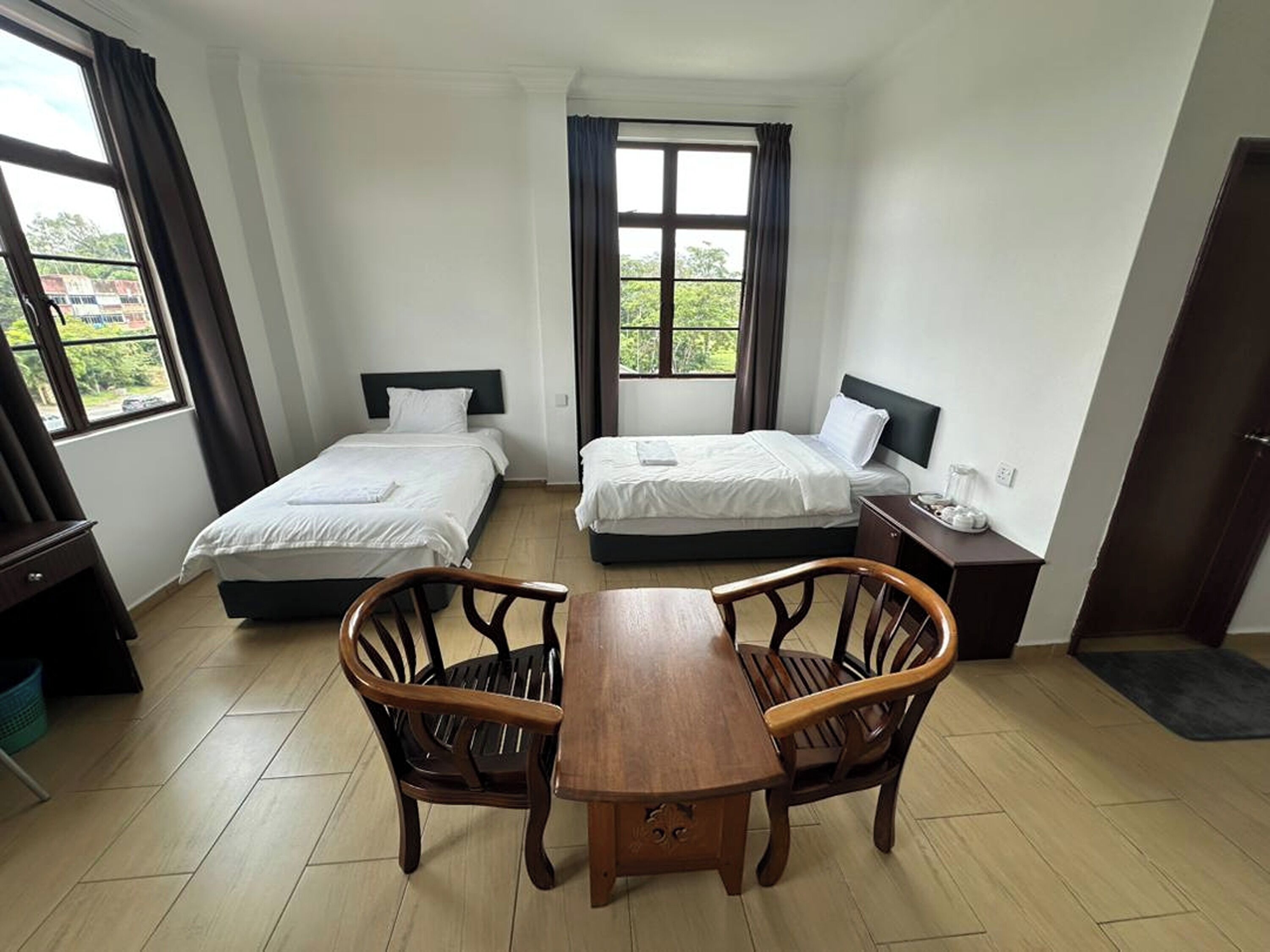 A hotel room with two single beds, a wooden dining table, and a view of greenery outside.