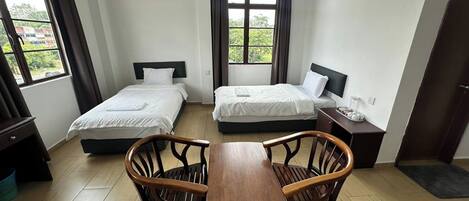 A hotel room with two single beds, a wooden dining table, and a view of greenery outside.