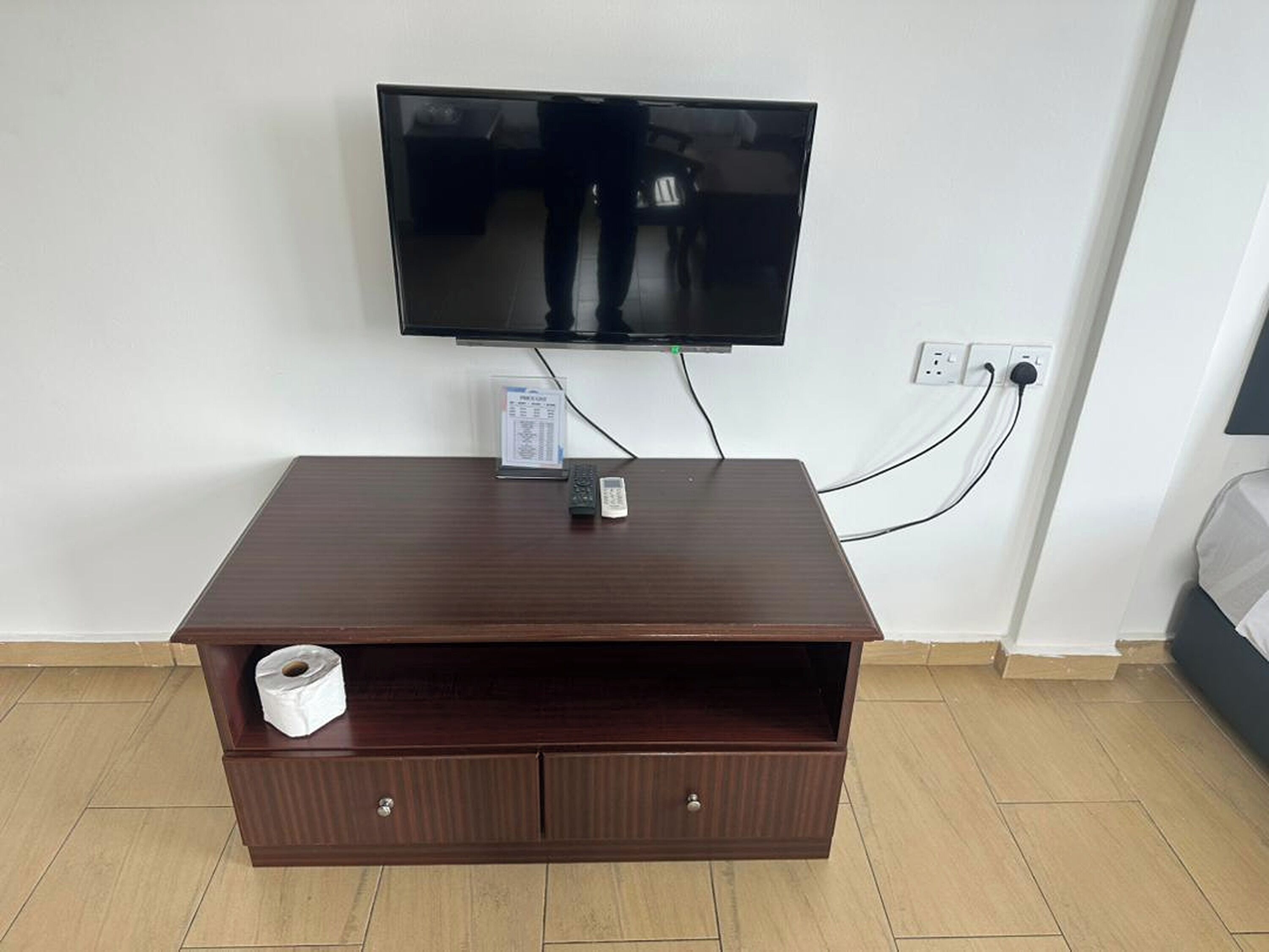 A hotel room with a flat-screen TV mounted on the wall, a wooden TV stand with a remote control, and a roll of toilet paper.