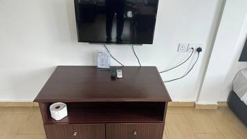 A hotel room with a flat-screen TV mounted on the wall, a wooden TV stand with a remote control, and a roll of toilet paper.